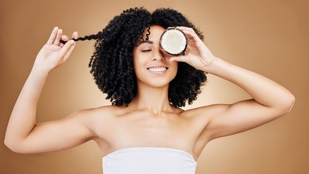 mulher negra com cabelos cacheados volumosos e saudáveis sorri enquanto segura uma metade de coco em frente ao olho.