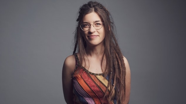 Mulher com óculos e dreads, vestindo blusa colorida listrada.