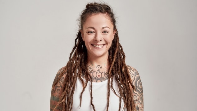 Mulher sorridente com dreads, tatuagens e regata branca.