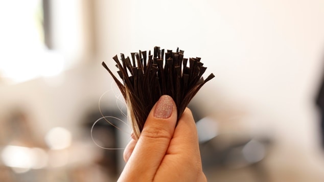 Mão com unhas delicadas segura um aplique de mega hair castanho com adesivo, pronto para ser aplicado.