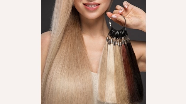 Mulher sorridente com longos cabelos loiros, segurando um mostruário circular com diversas mechas de mega hair em diferentes tons.