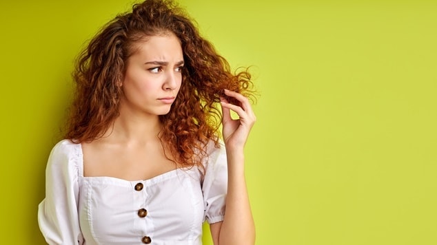 Pessoa com cabelo ondulado examina as pontas do cabelo, que aparentam estar ressecadas e com pontas duplas.