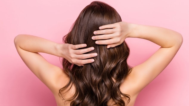 Mulher morena com cabelo ondulado colocando as mãos no seu couro cabeludo em um fundo rosa