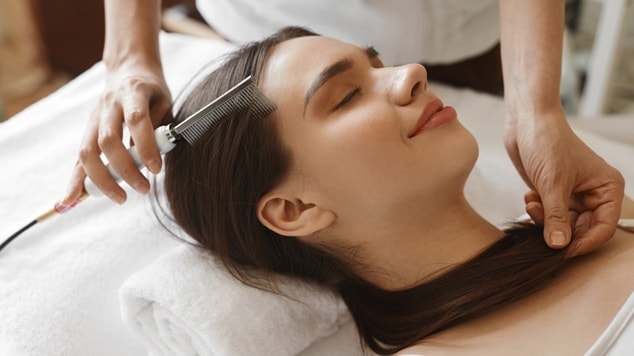 Mulher deitada na maca recebendo terapia capilar com um equipamento de um profissional.