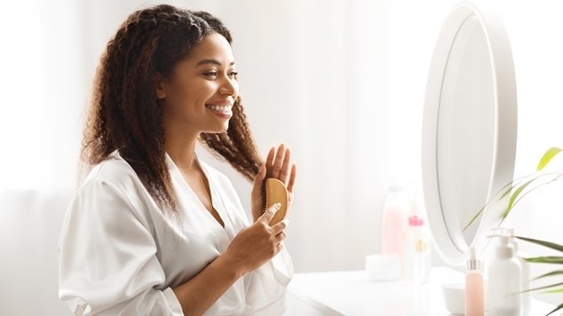  Mulher negra sorridente penteando seus cabelos cacheados com uma escova polvo, em frente ao espelho do banheiro.