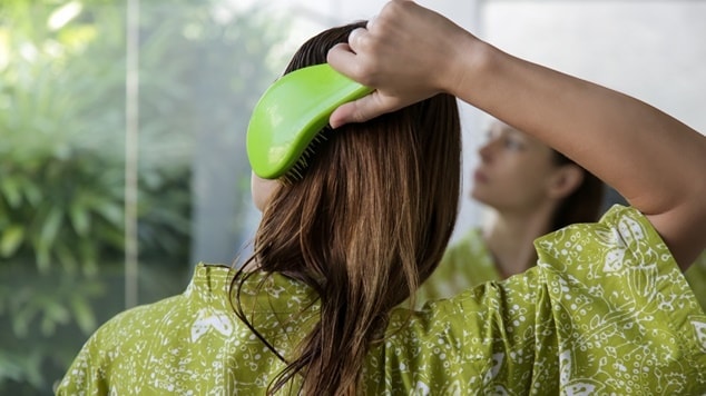 Vista traseira de uma mulher de roupão verde usando uma escova polvo verde para pentear seus cabelos úmidos.