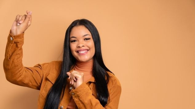 Mulher com cabelo liso, casaco laranja, fazendo um gesto de coração com os dedos e sorrindo.