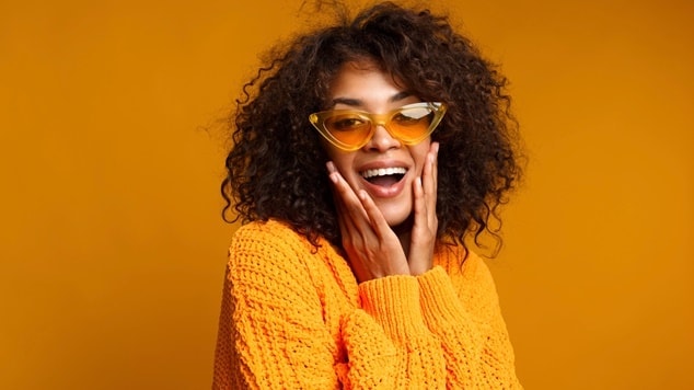 Mulher sorrindo com cabelo cacheado e óculos amarelos, vestindo um suéter laranja, em um fundo laranja.