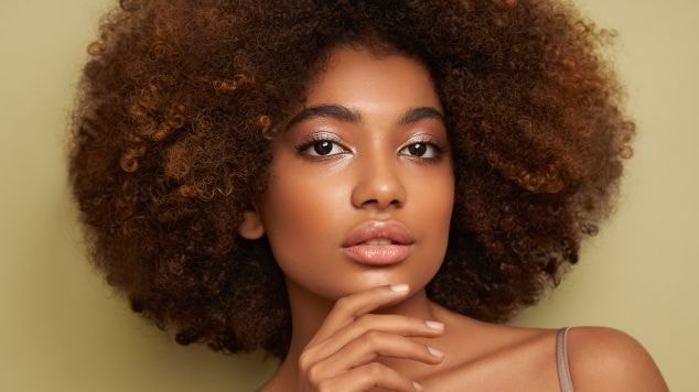 Mulher com mão no queixo, sombra perolada nos olhos e cabelo crespo na cor loiro escuro mel.