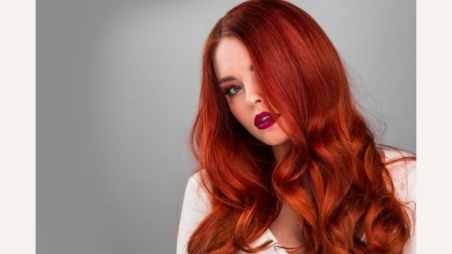 Mulher com cabelo ruivo intenso ondulado, marcante e  elegante em fundo cinza.