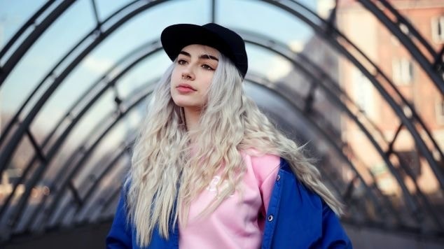 Jovem com cabelo platinado e ondulado em um cenário urbano, com estilo moderno e jovial.