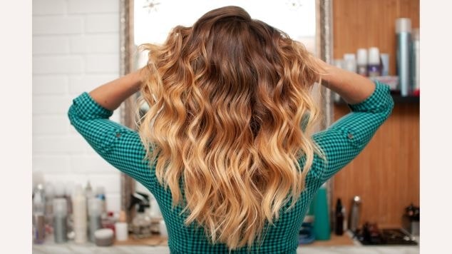 Mulher de costas com blusa azul com quadrados pretos segurando cabelo com ombre hair em tons de castanho e loiro.