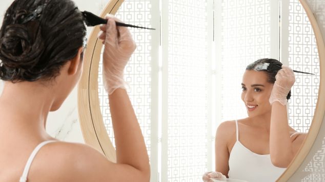 Mulher sorridente pintando o cabelo sozinha com luvas protetoras enquanto se olha no espelho. 