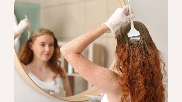 Mulher ruiva pintando o cabelo com luva enquanto olha no espelho.