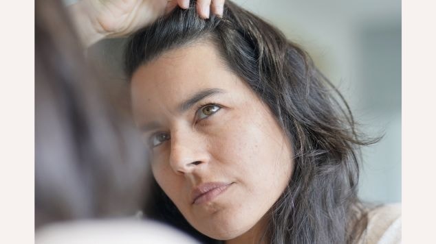 Mulher observa os fios brancos na raiz dos cabelos ao se olhar no espelho, avaliando o crescimento dos grisalhos.