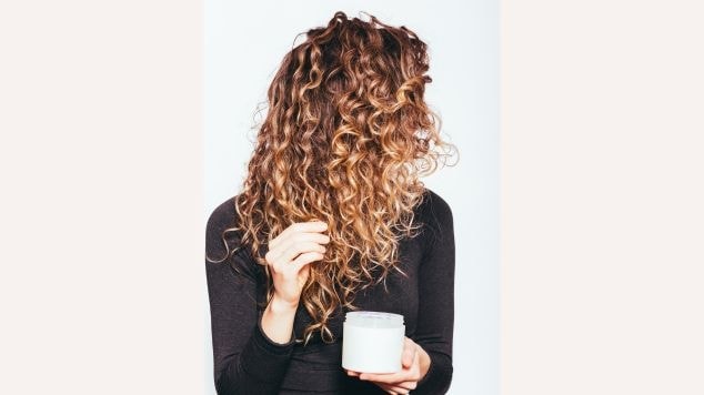Mulher de costas com cabelos cacheados em degradê de castanho para loiro segurando um pote branco de creme para cabelos enquanto toca seus cachos com a outra mão. Ela veste uma blusa preta e está contra um fundo branco.
