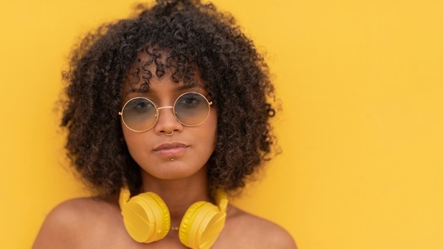 Mulher com fone de ouvido amarelo no pescoço, com cabelo ondulado e óculos de sol no rosto