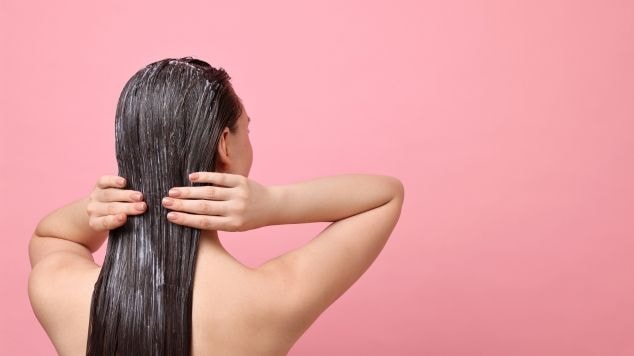 Mulher passando máscara no cabelo em fundo rosa.