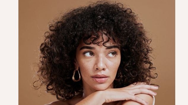 Mulher com cabelos cacheados bem definidos e hidratados exibindo cachos perfeitos em fundo marrom neutro que realça a textura e brilho dos fios.