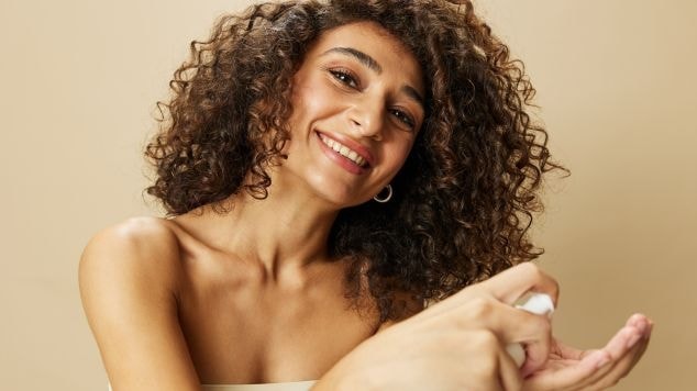 Mulher sorridente aplicando creme de pentear em seu cabelo cacheado natural, demonstrando a distribuição do produto com as mãos sobre o fundo bege.