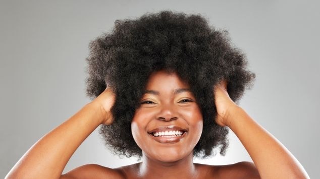 Mulher sorridente com cabelo crespo volumoso e bem definido colocando as duas mãos no seu cabelo.