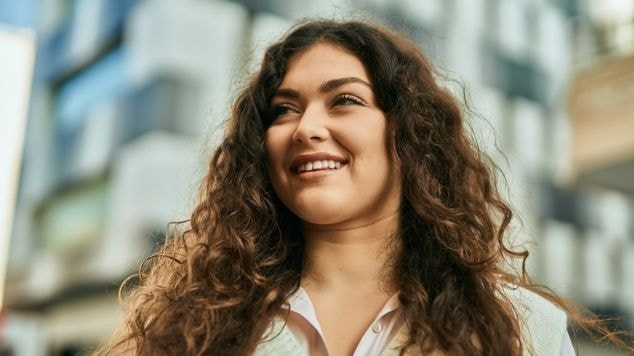 Mulher sorridente com cabelos ondulados hidratados e usando blusa em tom claro.
