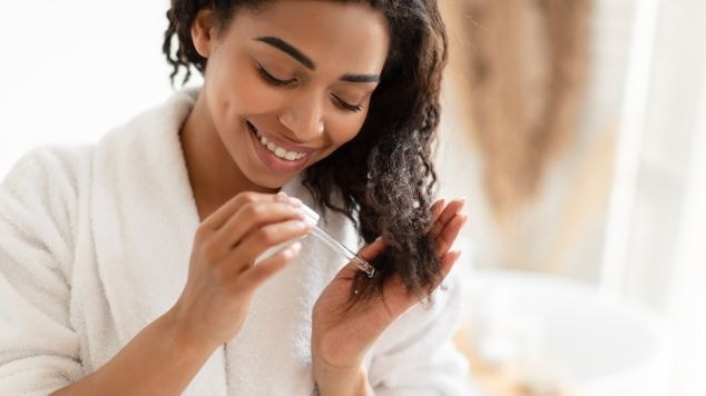 Mulher sorridente usando roupão branco aplicando produto capilar em seus cabelos cacheados, demonstrando cuidados com o cronograma capilar em casa.