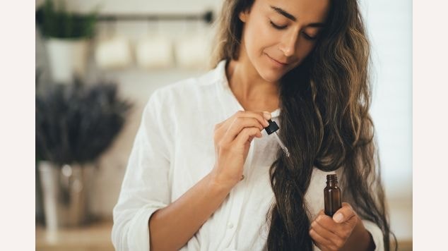 Mulher aplicando pré shampoo em óleo no seus cabelos longos ondulados.