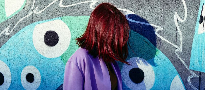 Mulher com cabelo marsala intenso em frente a um mural de grafiti azul.