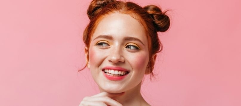 Mulher jovem de pele clara e cabelo ruivo vibrante penteado em dois coques, sorrindo enquanto olha para o lado