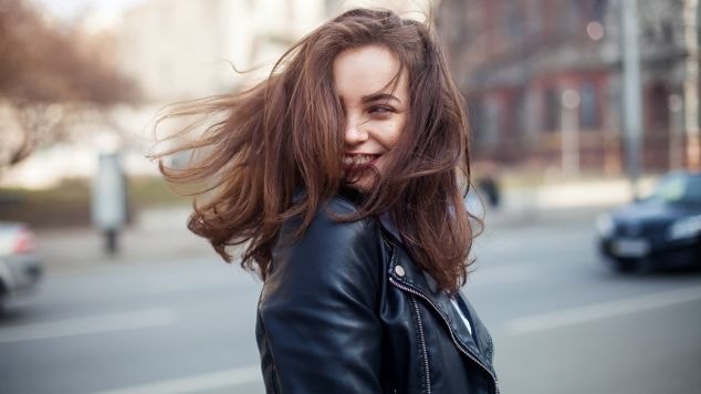 Mulher com cabelo castanho solto ao vento, jaqueta de couro preta, sorrindo na rua. Visual moderno, cheio de atitude e energia.
