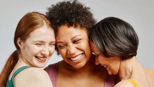 Três mulheres felizes com diferentes tipos de cabelo e colorações se abraçam, representando diversidade, beleza e cuidado capilar.