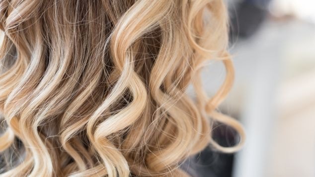 Fotografia com foco na parte anterior do cabelo ondulado com mechas loiras de uma mulher no salão de beleza.