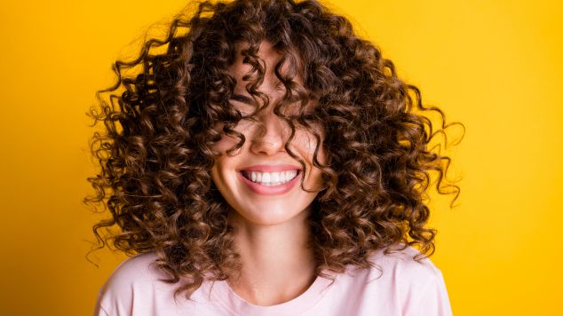 Jovem sorridente com o cabelo cacheado utiliza defrizante com proteção térmica para acabar com o frizz no cabelo.