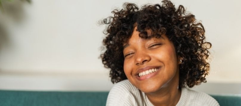 Mulher jovem de cabelos cacheados curtos sorrindo de olhos fechados. Seus cabelos estão naturalmente brilhantes e saudáveis, exibindo os efeitos da biotina e vitamina B5 no cabelo.