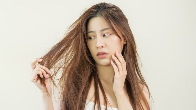 Jovem modelo em fundo branco analisa seu cabelo castanho liso e danificado e considera realizar uma blindagem nos cabelos para protegê-los.