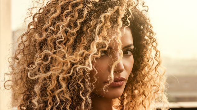Mulher com cabelo cacheado volumoso e definido, com mechas loiras e castanhas, exibindo cachos saudáveis e brilhantes em luz natural dourada ao entardecer.