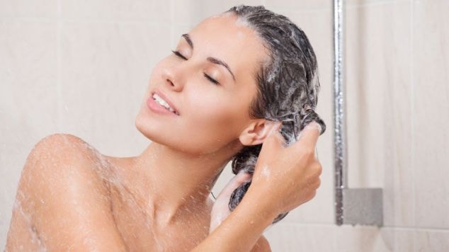 Mulher lavando o cabelo no chuveiro, massageando os fios com shampoo e os olhos fechados, demonstrando o momento de cuidado durante a lavagem capilar.