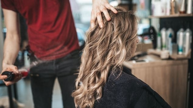 Cabeleireiro estiliza cabelo ondulado e loiro em salão de beleza, com produtos capilares ao fundo.