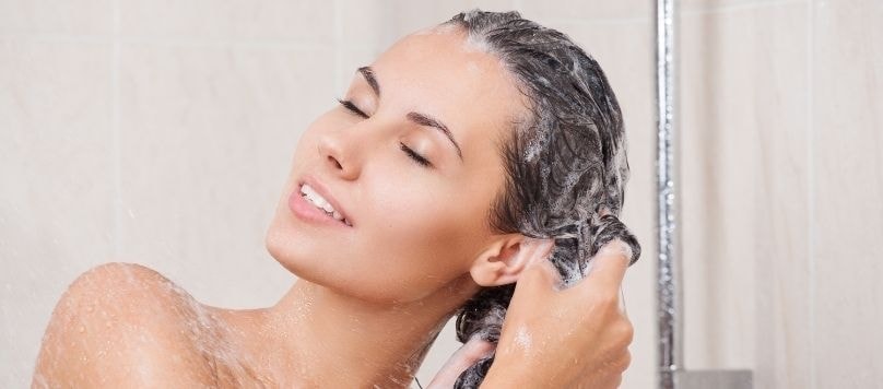 Mulher branca enxaguando os fios ensaboados do cabelo no chuveiro após fazer um detox capilar.