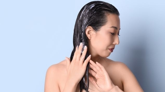 Mulher aplicando máscara capilar nos fios molhados, massageando o couro cabeludo com as mãos durante o tratamento capilar no banho.