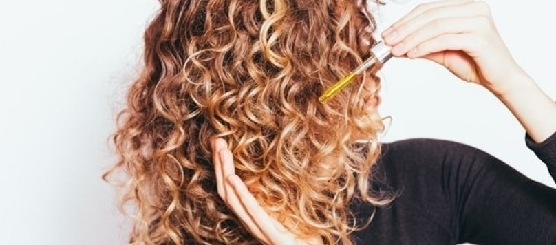 Mulher cacheada com cabelos castanhos com pontas loiras descoloridas aplicando óleo de argan com uma pipeta no comprimento de seus cabelos.