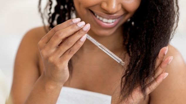 Mulher sorrindo enrolada na toalha aplicando produto nas pontas dos cabelos com um conta gotas.
