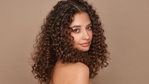 Mulher de cabelos cacheados volumosos e definidos, olhando por sobre o ombro, com fundo neutro em tom nude.