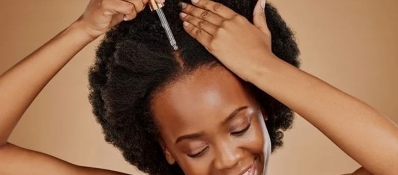 Mulher negra com cabelos crespos fazendo umectação no cabelo, aplicando óleo capilar na raiz com uma pipeta e com as mãos.
