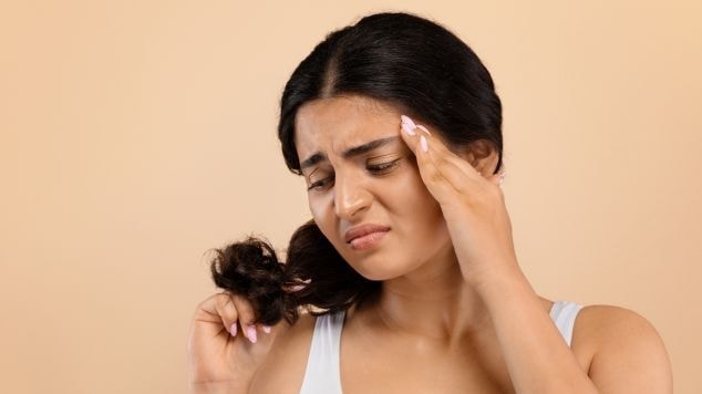 Mulher de regata branca com a mão apoiada na têmpora olhando para o cabelo com cara de preocupação em um fundo bege.
