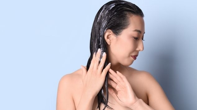 Mulher no chuveiro com o cabelo molhado colocando shampoo no cabelo com as duas mãos.