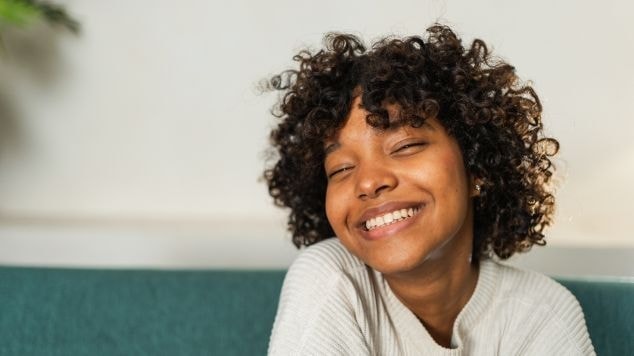 Mulher jovem de cabelos cacheados sorri espontaneamente, olhos fechados, sentada em um sofá verde-água, usando suéter branco de tricô.