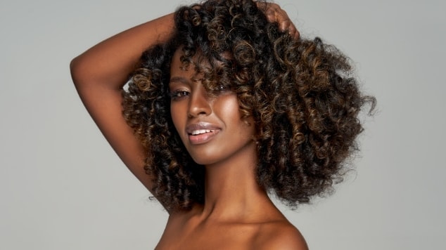Foto de uma mulher negra esticando uma mecha de seu cabelo para demonstrar a curvatura dos cachos.