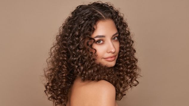 Mulher jovem de cabelos cacheados castanhos longos e definidos, olhando para a câmera com leve sorriso, usando top preto em fundo bege neutro.